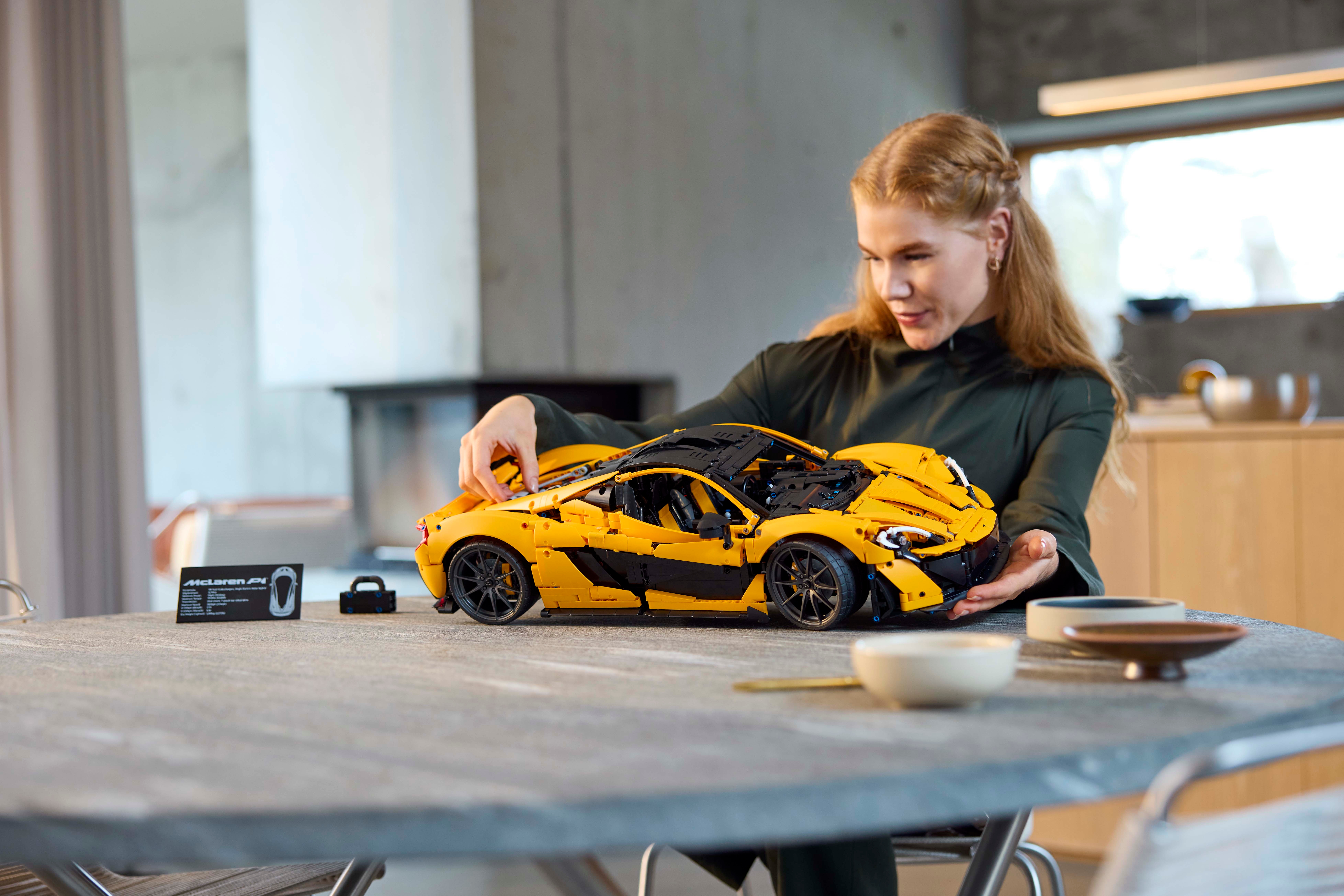 Lifestyle image of a woman with long hair sitting by a table with the new LEGO Technic McLaren P1 build infront of her while she is admiring the finished model