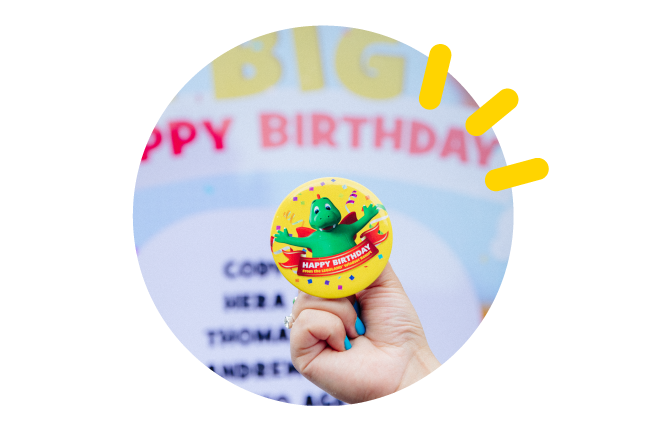 A picture bubble of a hand holding a badge featuring a dinosaur and the text "Happy Birthday!"